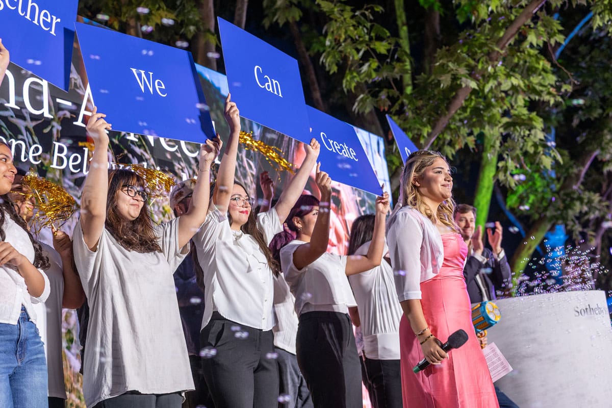 A young woman in a pink dress holding a microphone. Behind her, a group holding up blue signs reading "together we can create..."